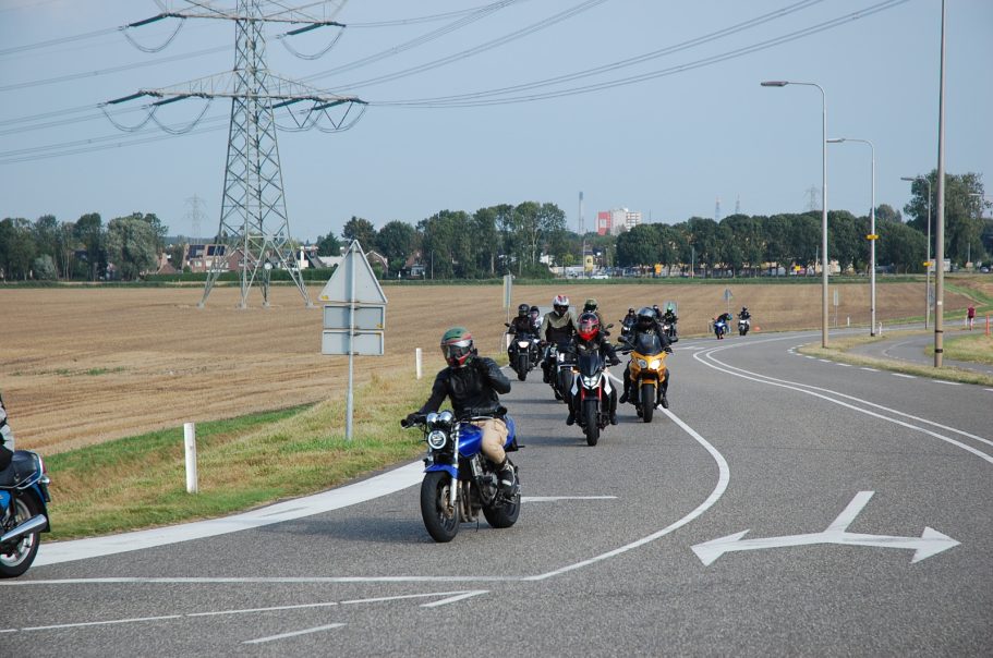 Groep motorrijders rijdt op een landelijke weg met elektrische masten op de achtergrond.